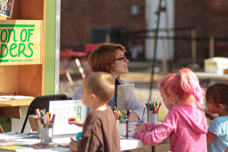 Three children draw at a table outside while a person smiles into the distance