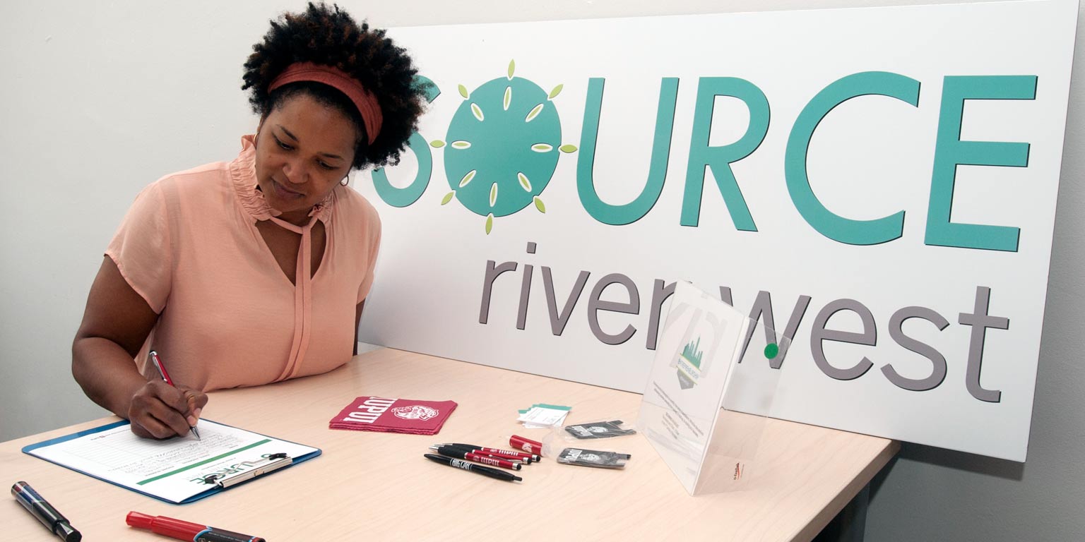 A person fills out a form at a table with a sign that says Source River West.